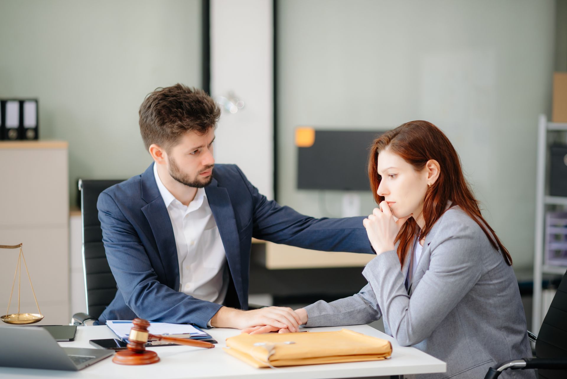 Two coworkers in an office, one comforting the other at a desk with folders and a gavel.