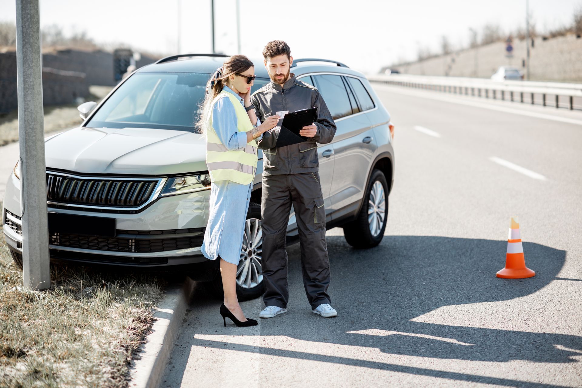 Two roadside workers inspecting a black SUV on the shoulder beside an orange cone
