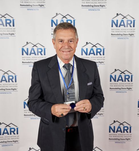 Man in suit smiling, holding a blue badge in front of a blue and white NARI backdrop.