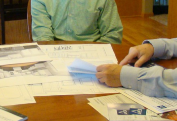Two men at a table reviewing blueprints and documents; one points at paper, other is seated.
