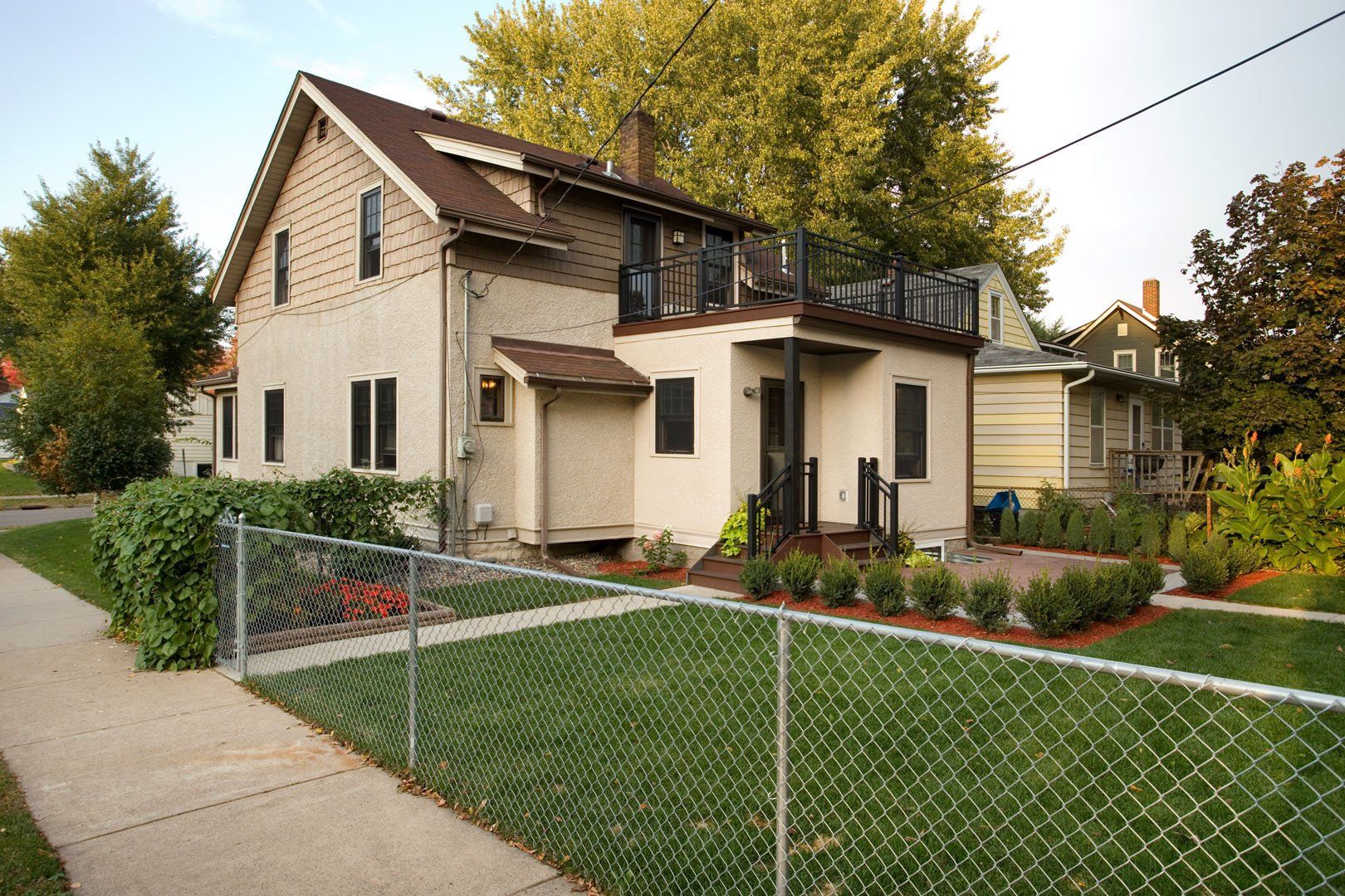 Two-story tan house with a fenced-in yard and rooftop deck; trees in the background.