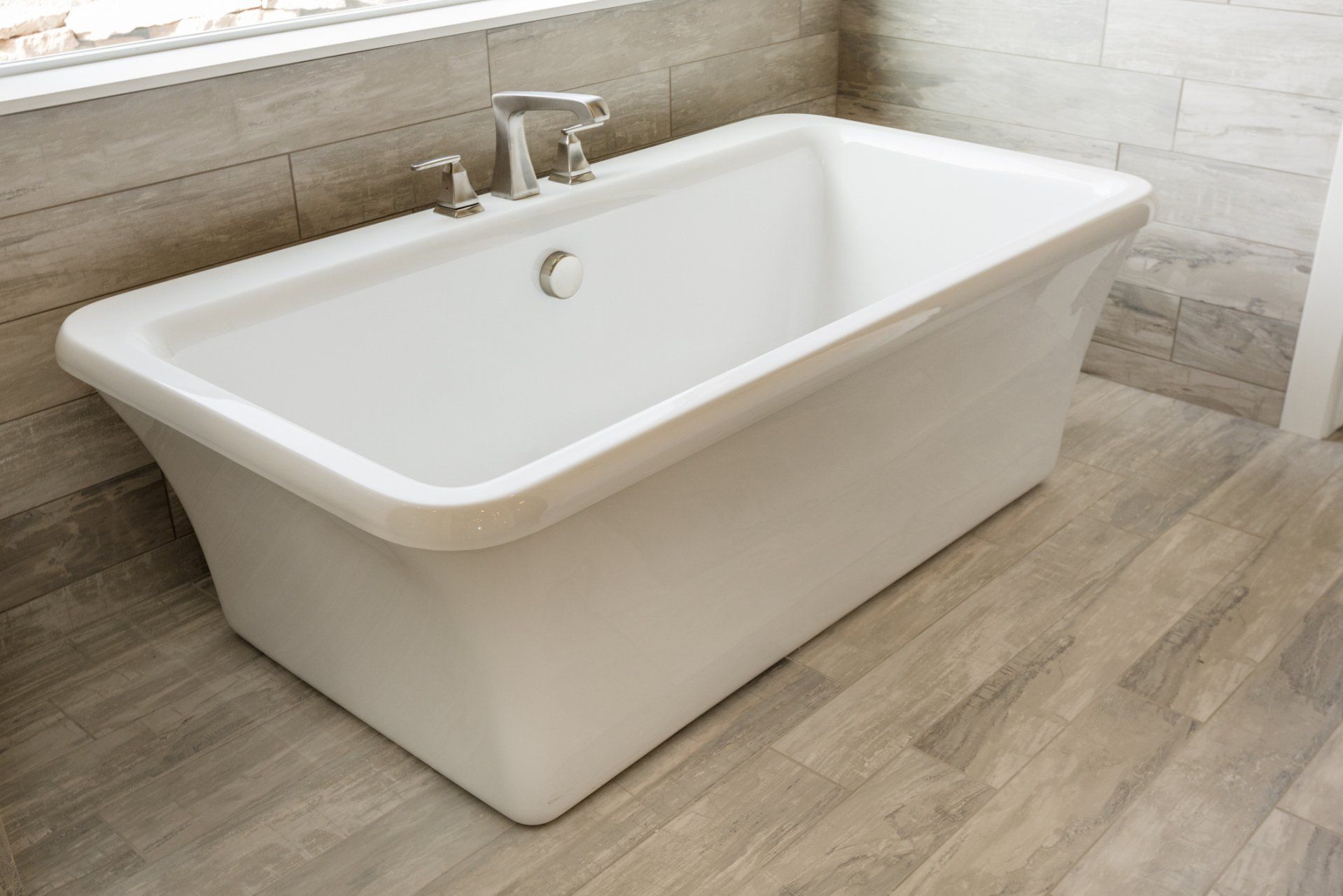 White rectangular bathtub in a modern bathroom with light-colored tile walls and flooring; chrome faucet.