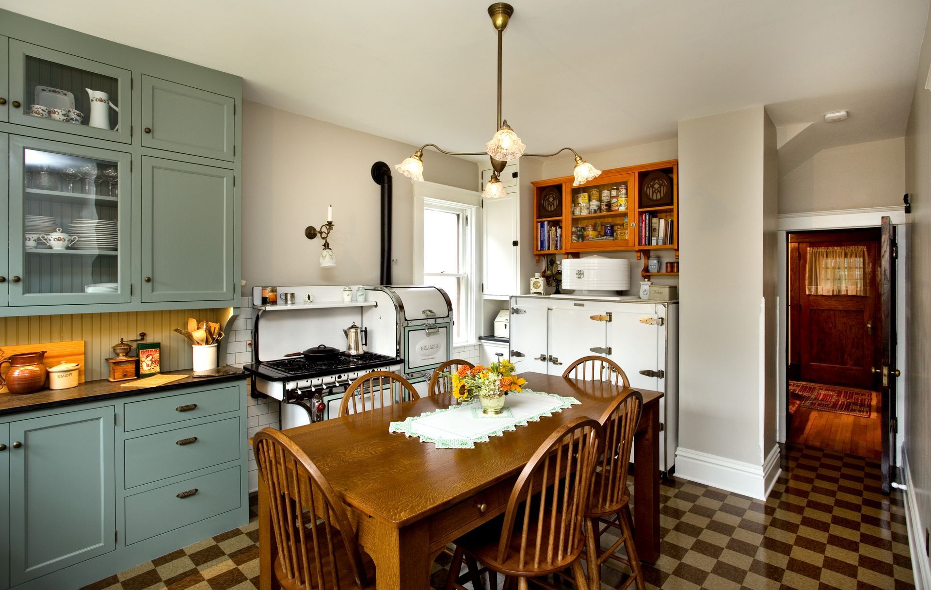 A vintage kitchen with green cabinets, a wooden table, and a black and white tiled floor.