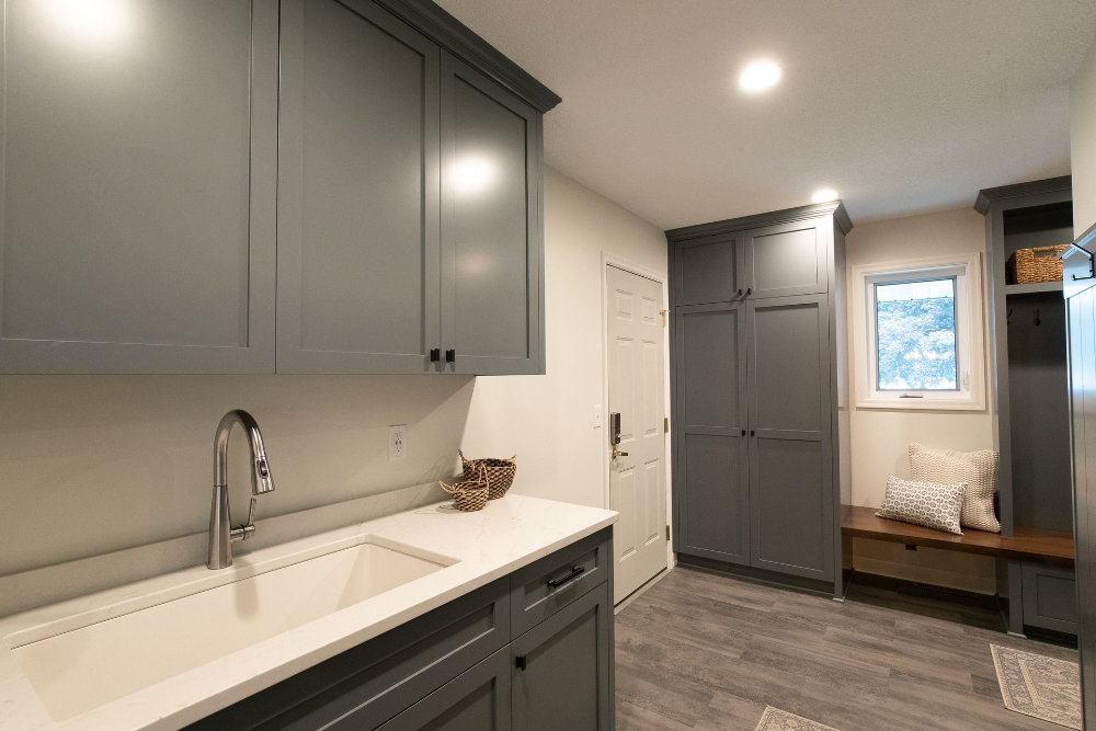 Gray laundry room with white countertop, cabinets, and a sink. Door and bench.