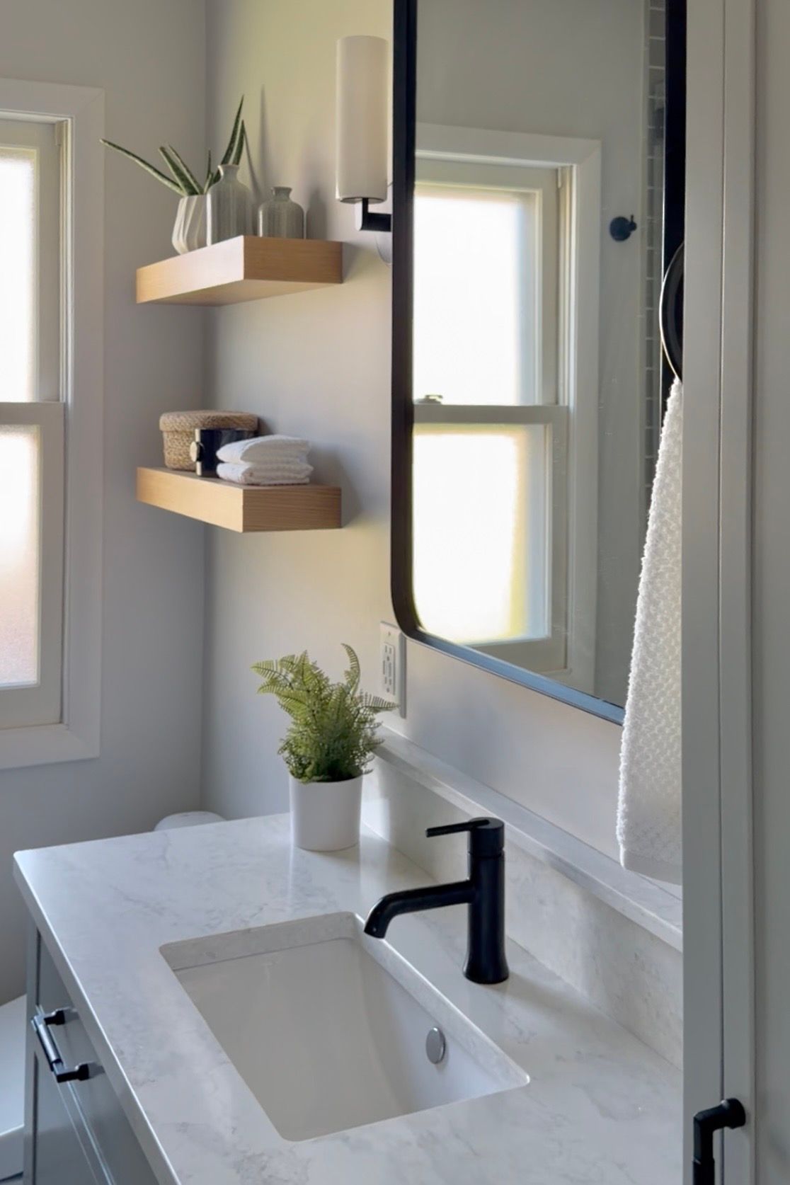 Modern bathroom vanity with white sink, black faucet, mirror, small plant, and wooden shelves by a window