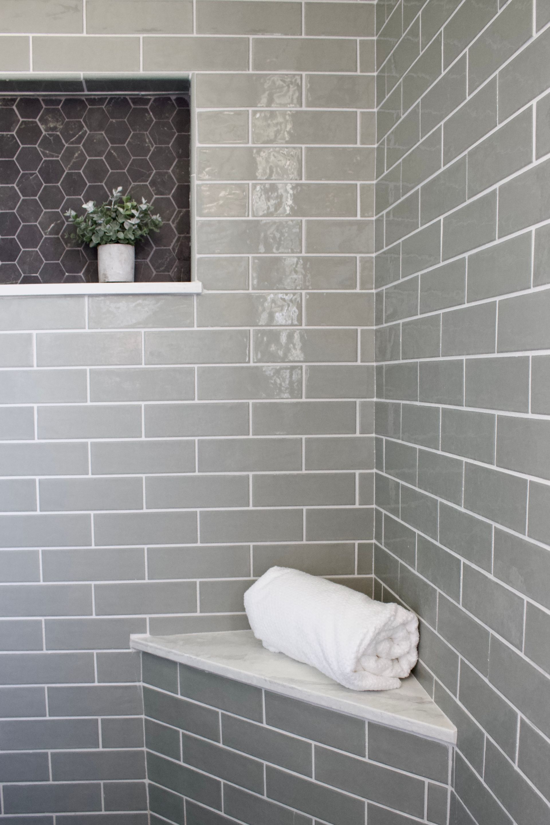 Gray tiled shower with a corner bench, white towel, and small potted plant on a recessed shelf.