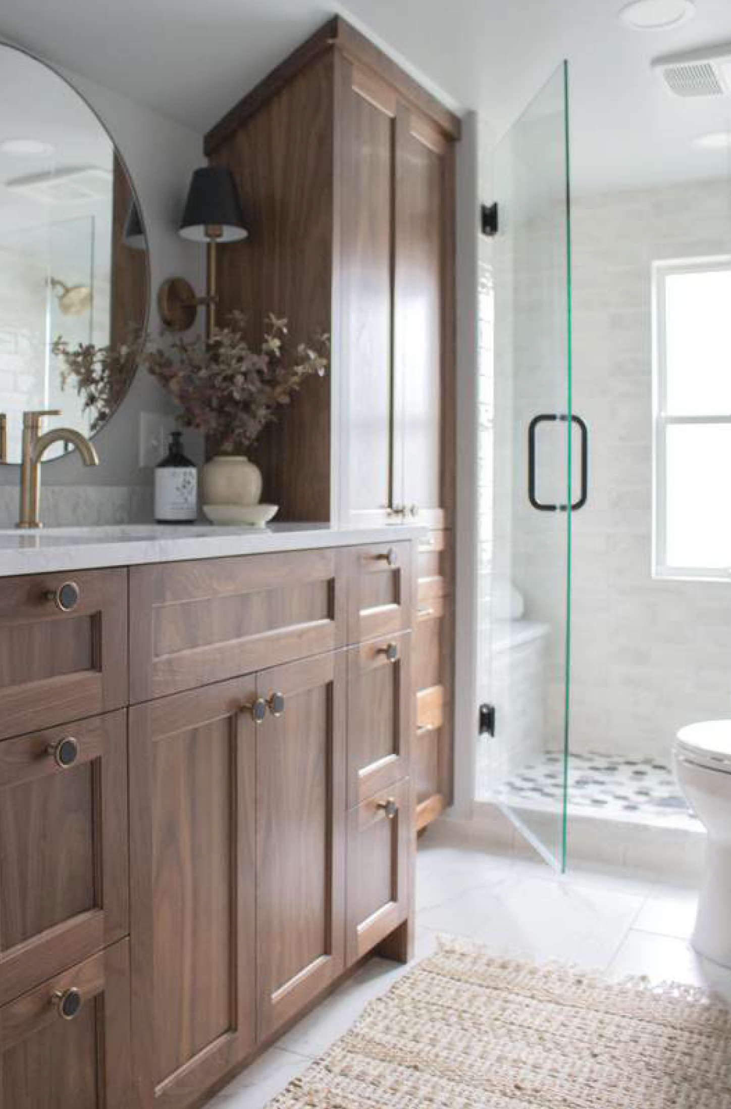 Bathroom with wood cabinets, glass shower, round mirror, white tile, and a neutral rug.
