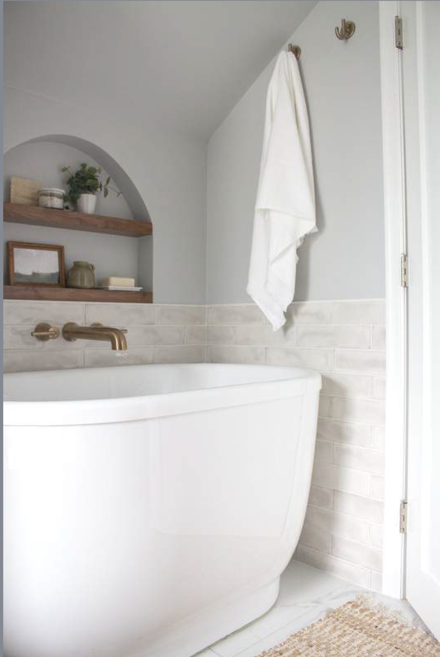 Bathroom with white tub, light gray walls, wooden shelves with decor, and a towel hanging.