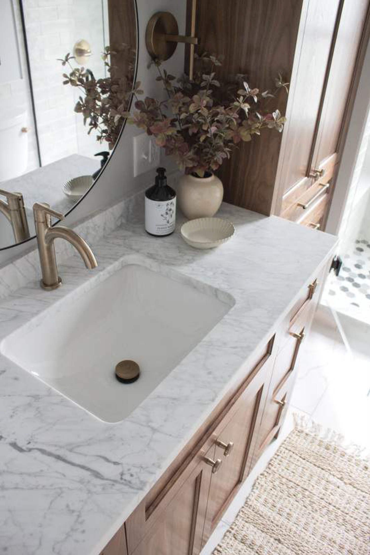 Bathroom vanity with marble countertop, gold faucet, and wooden cabinets. Dried flowers in a vase and soap dispenser.