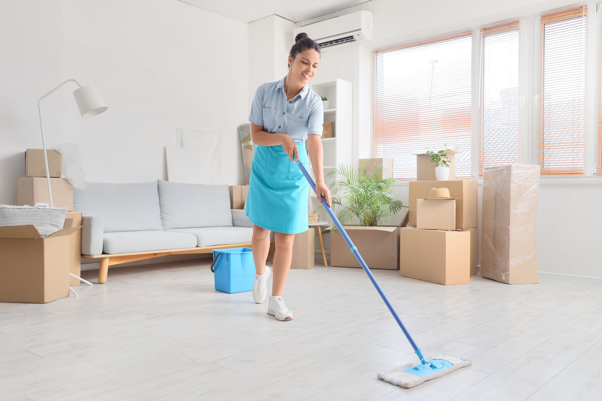 Woman mopping a white floor in a room with moving boxes, a sofa, and large windows.