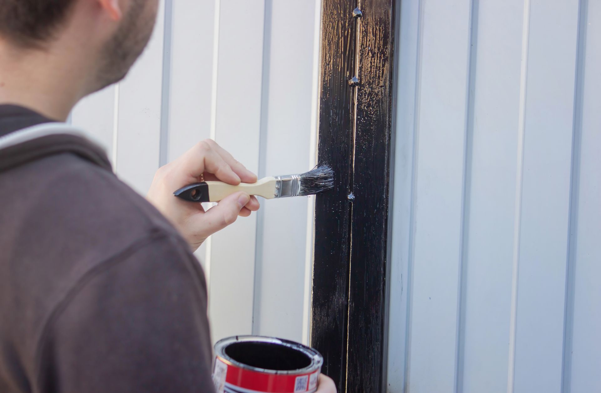 Person painting a black post on a white fence with a paintbrush and a can of paint.