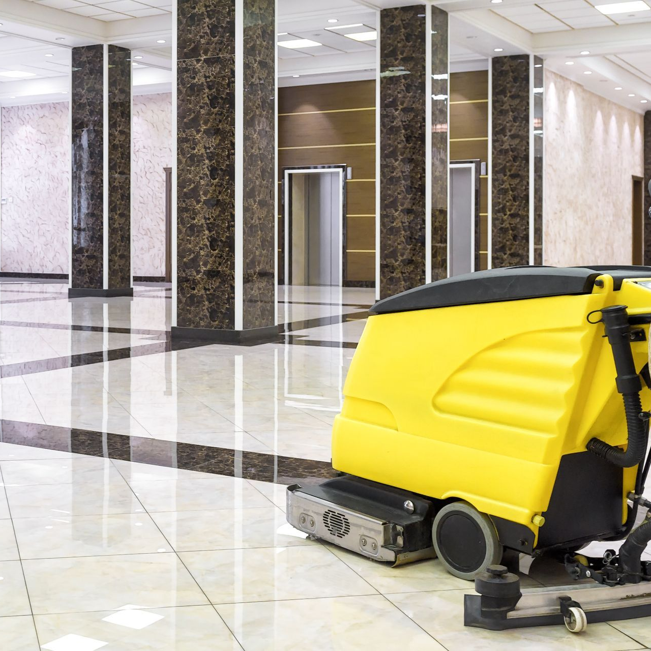 Yellow floor cleaner in a polished marble-floored lobby, cleaning. Columns and elevators in the background.
