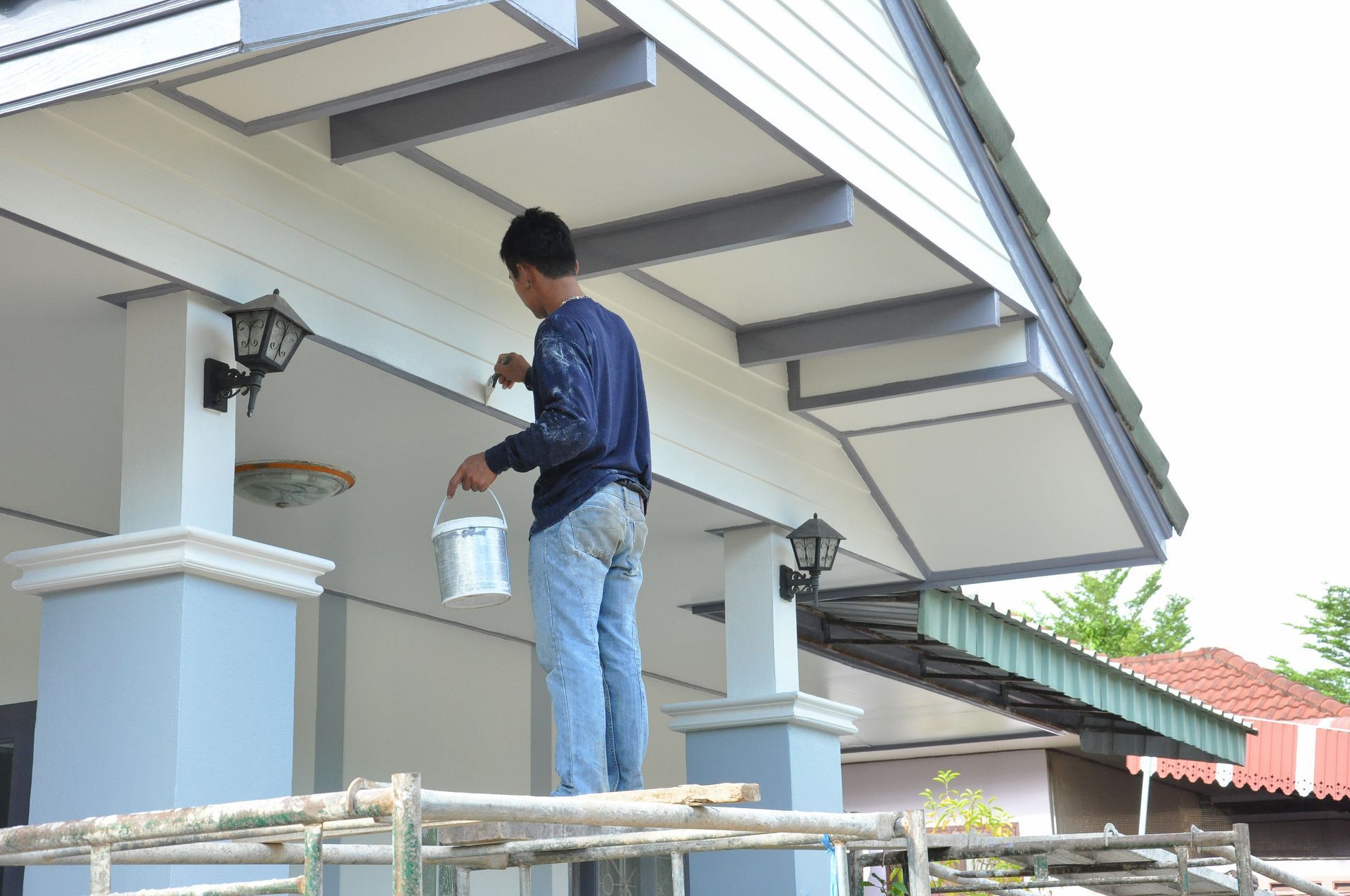 Person painting a building's exterior. The man stands on scaffolding, applying white paint to the structure's light blue trim.