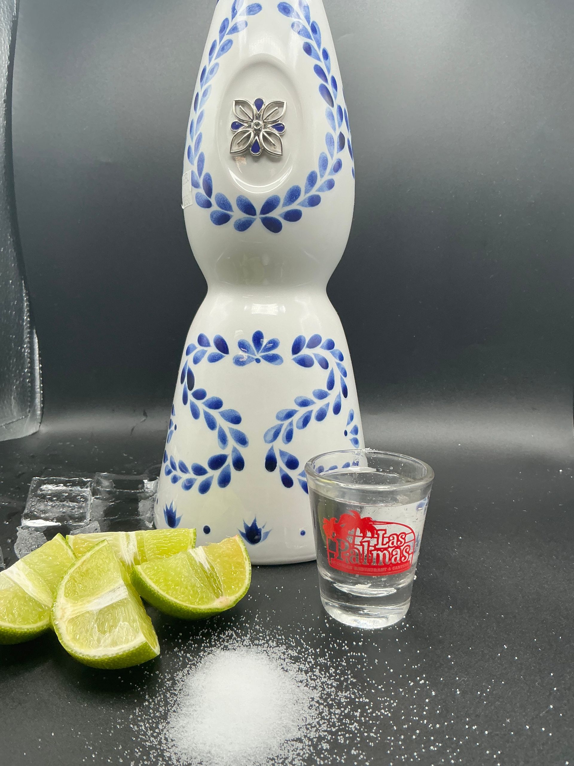 Tequila bottle, shot glass, limes, salt, and ice on a black surface.