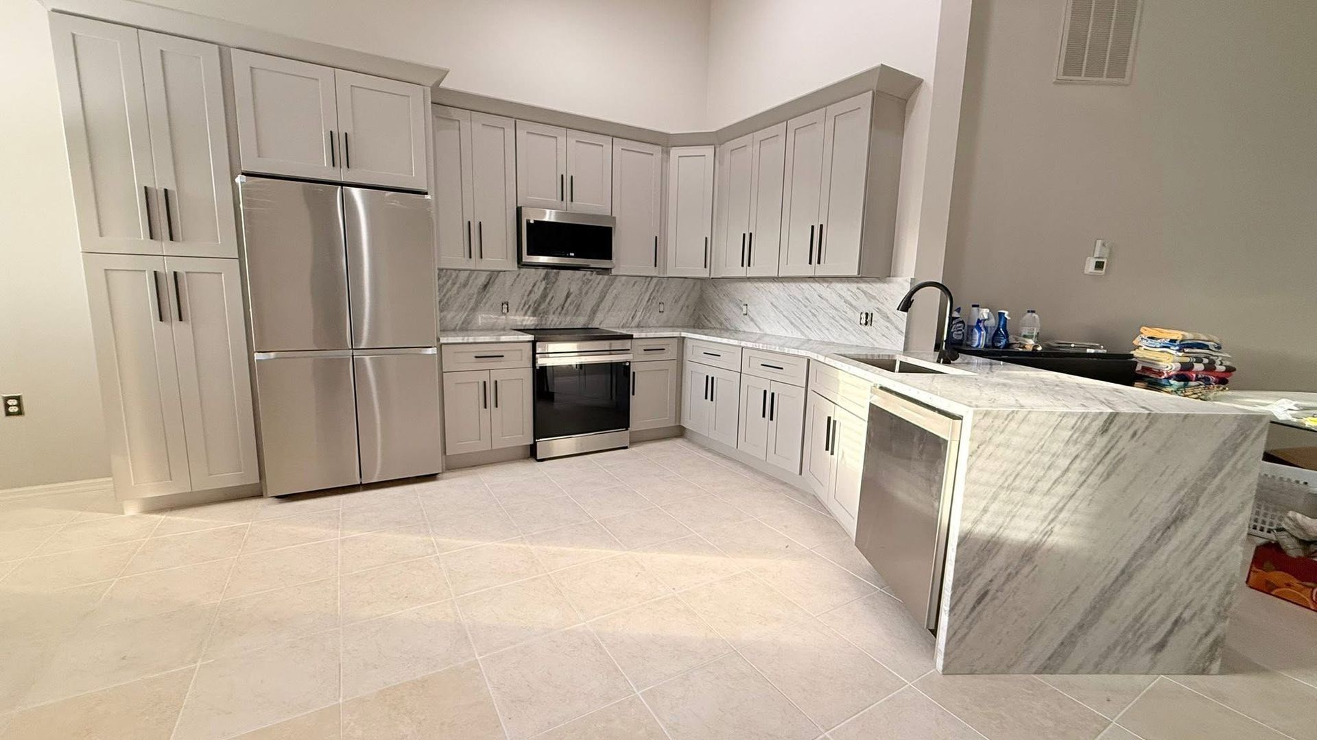 Modern white kitchen with stainless steel appliances, marble island, and pale tile floor.
