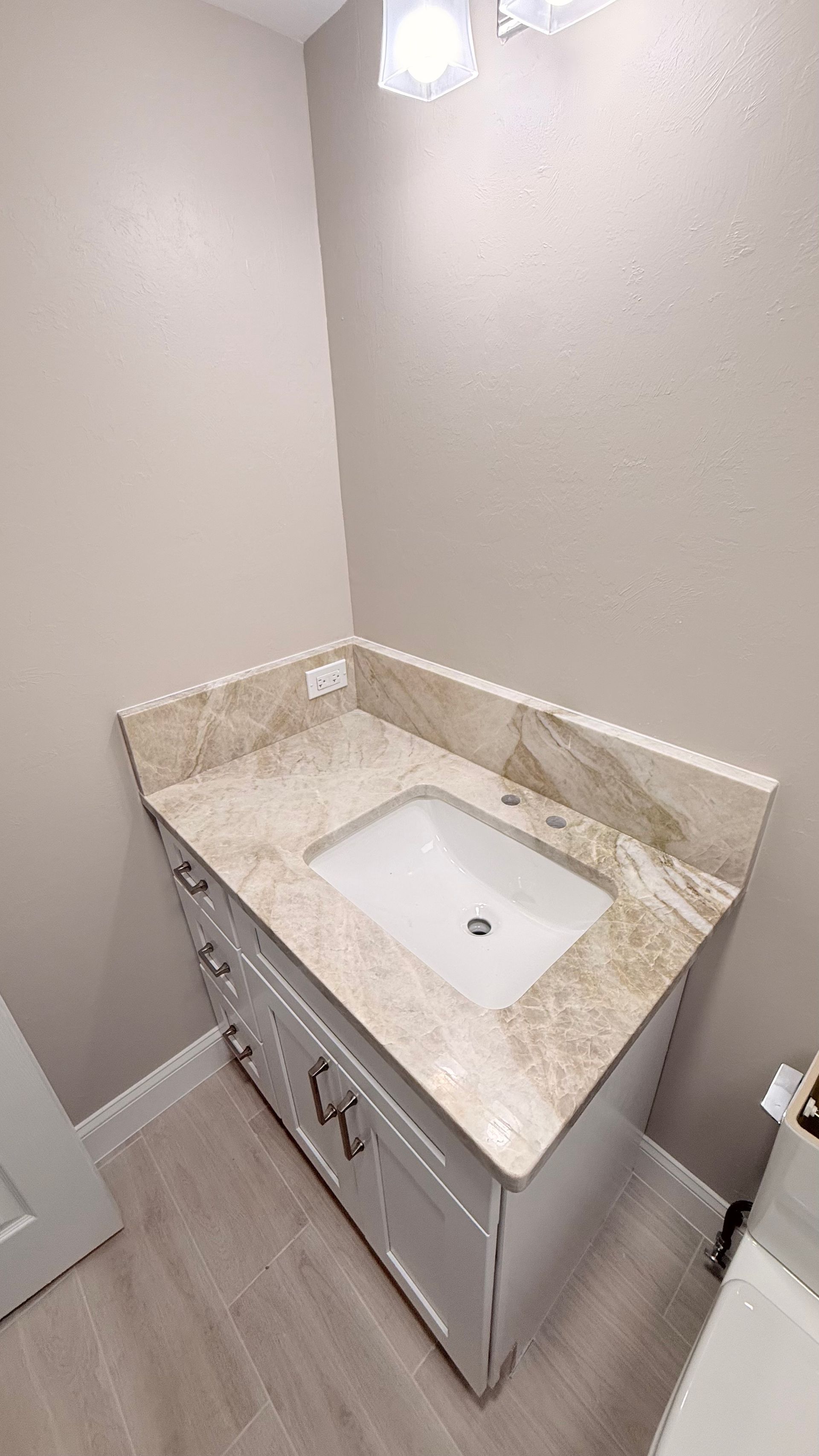 Small bathroom vanity with white sink, beige marble countertop, and gray cabinets under a wall light.