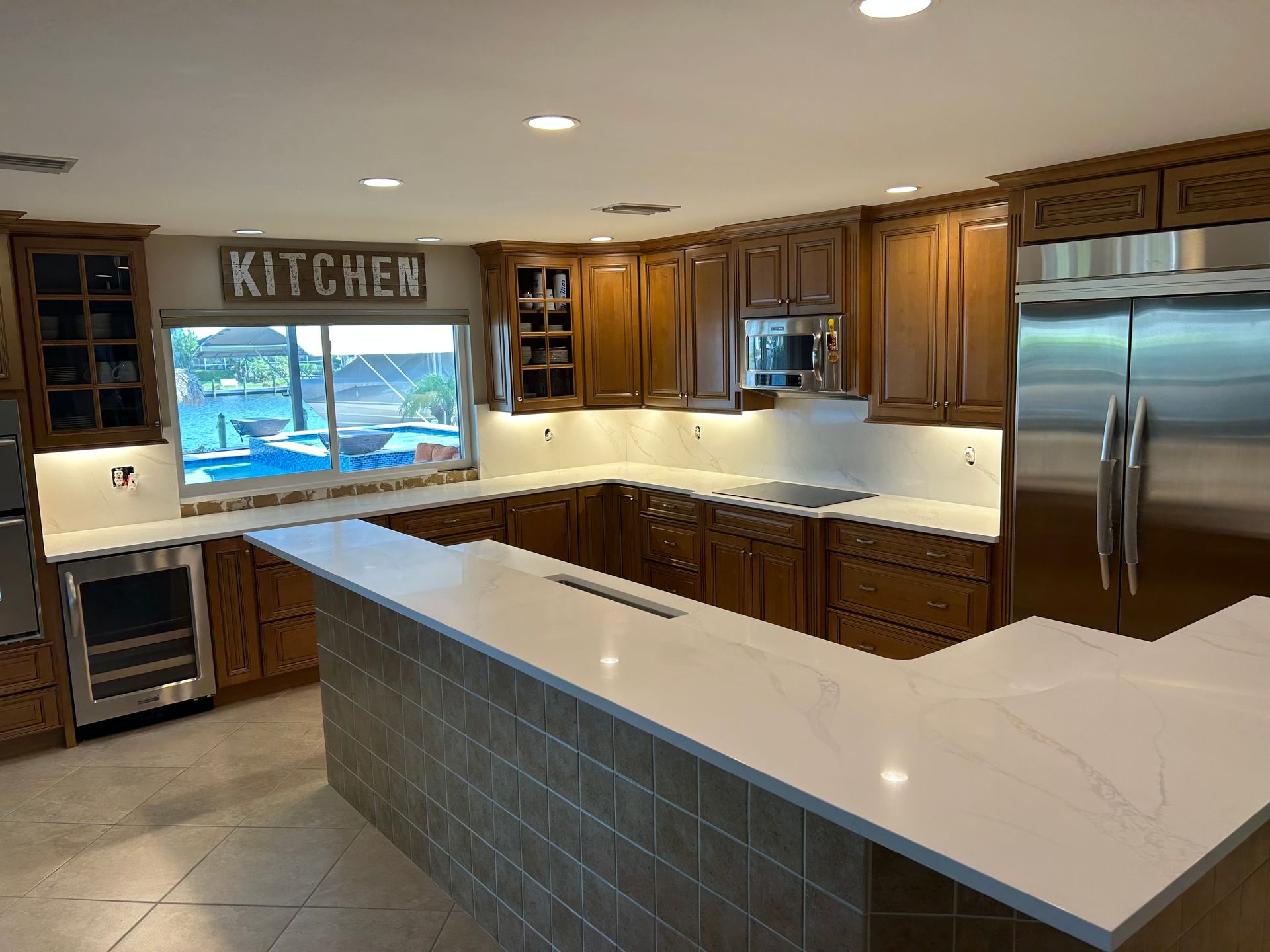 Modern kitchen with white countertops, wood cabinets, stainless steel appliances, and a large window over the sink