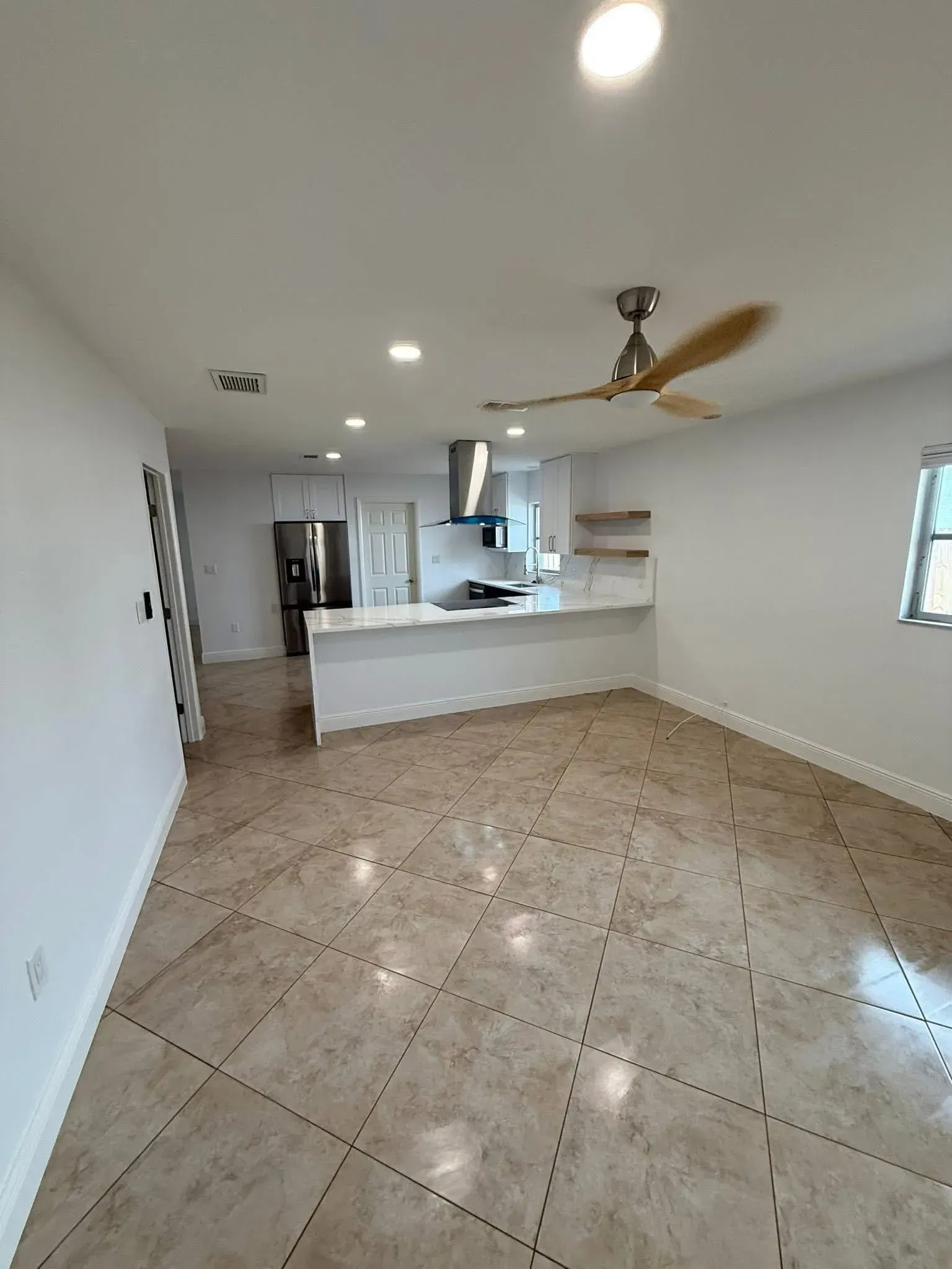 Empty tiled kitchen and dining area with white walls, island counter, and ceiling fan under recessed lights
