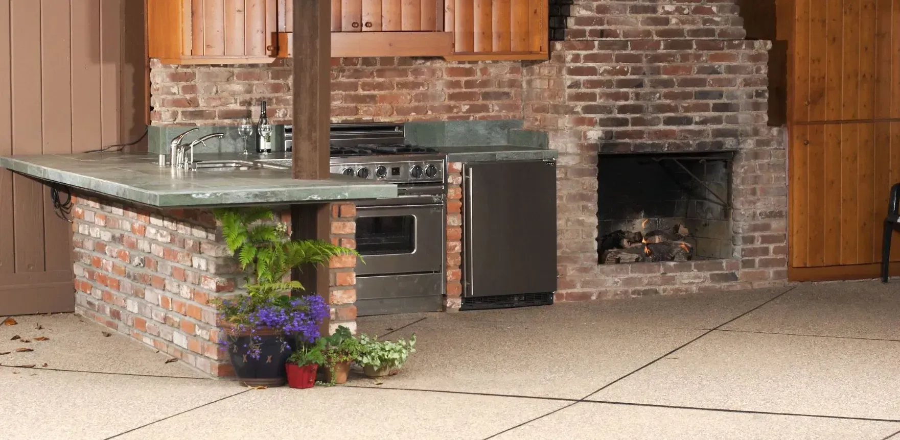 Indoor kitchen with stone island, brick grill, and wood cabinets, with a small potted plant on the floor.