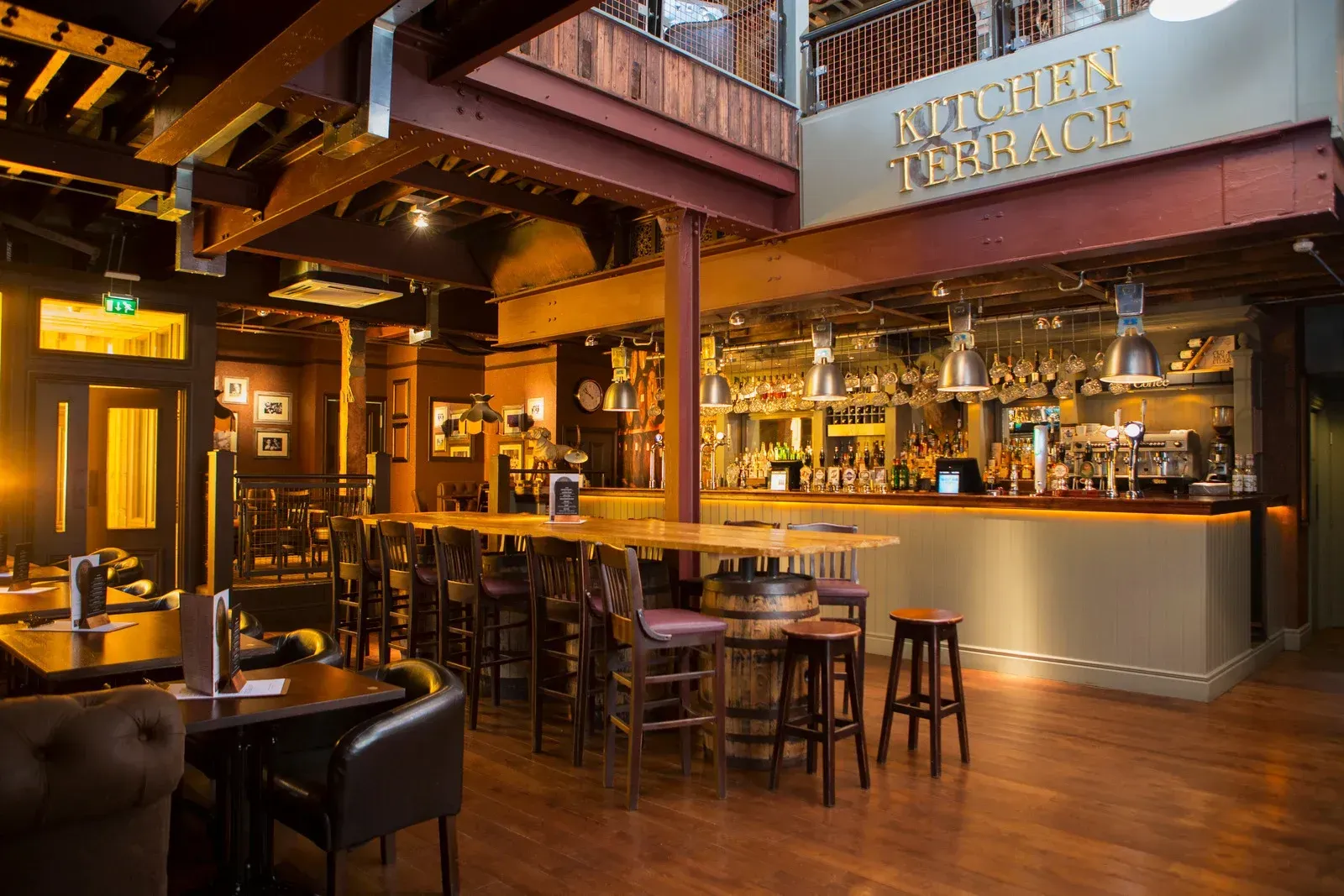 Warmly lit restaurant bar with wooden booths, high stools, and a glowing counter under a loft balcony.