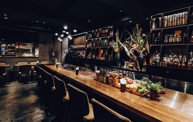 Dimly lit bar interior with a long wooden counter, stools, and shelves of liquor bottles.