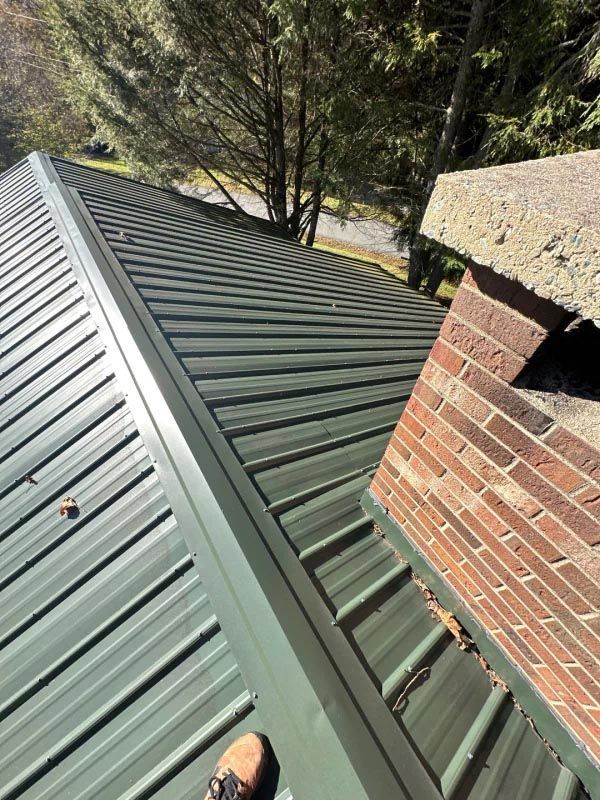 A person is standing on a green metal roof next to a brick chimney.