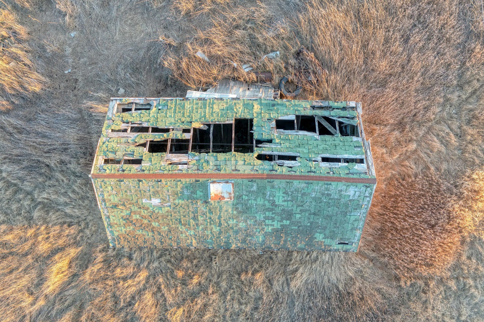 Aerial view of a weathered, rectangular green building with a severely damaged roof surrounded by dry, brownish grass.