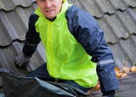 A person wearing a high-visibility yellow and navy rain jacket, kneeling on a dark tiled roof.