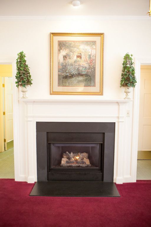 Fireplace with painting above, topped with potted greenery. Red carpet, white walls.