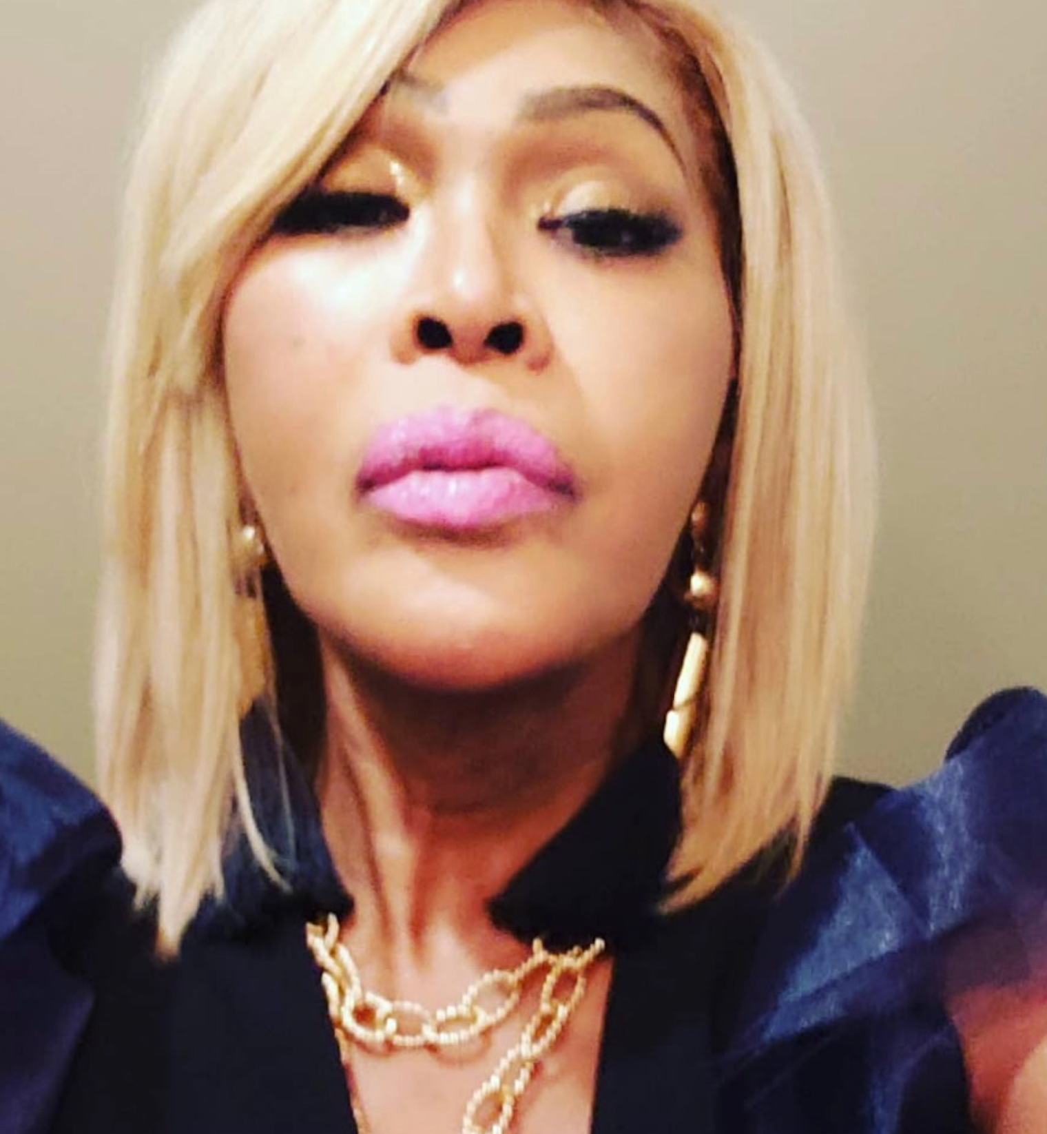 A close-up selfie of a person with blonde hair, bright pink lipstick, and a gold chain necklace, wearing a dark top.