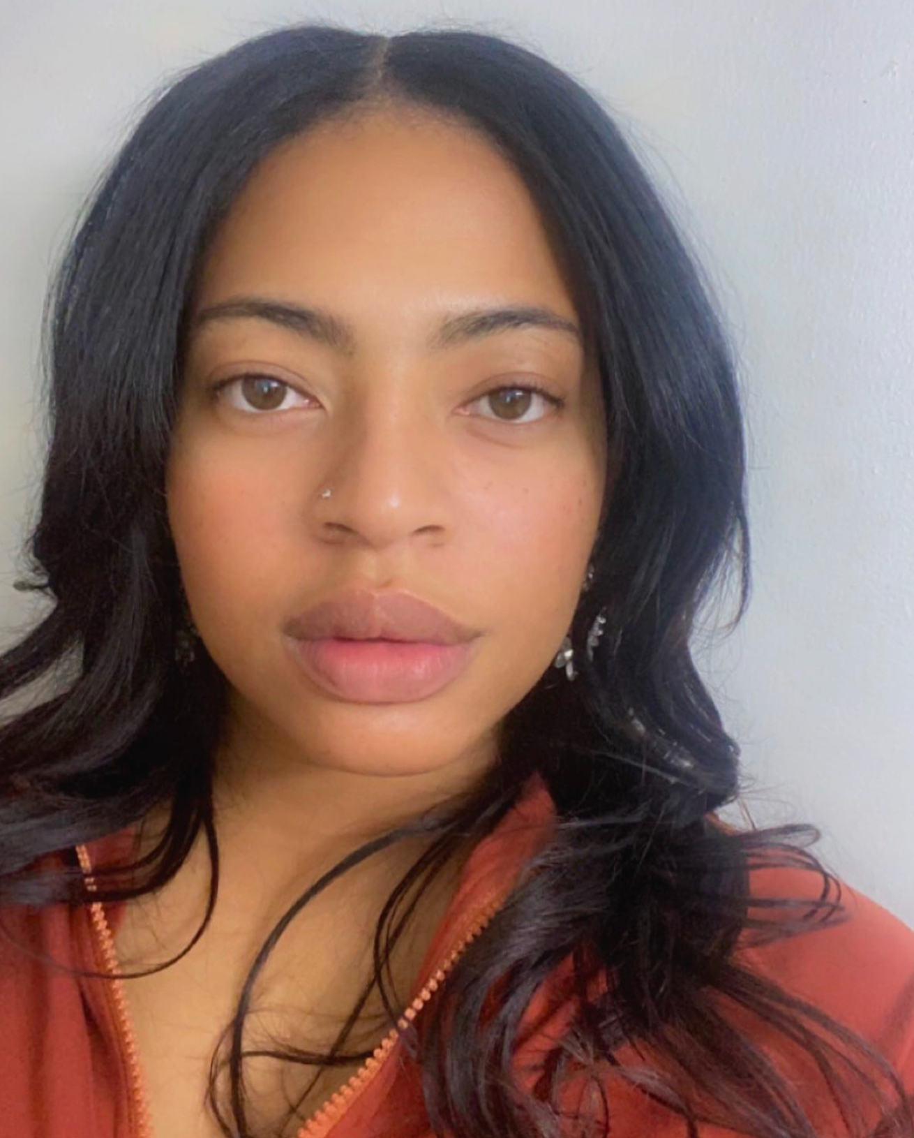 A portrait of a person with long, dark, wavy hair and a nose ring, wearing a rust-colored jacket against a plain wall.