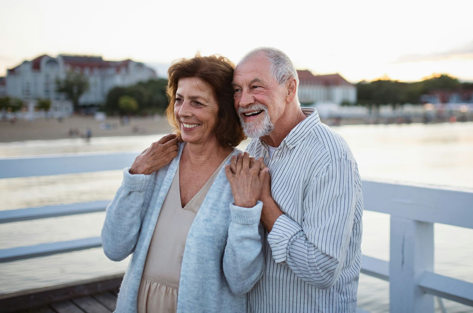 Smiling elderly couple on a wooden pier, overlooking the ocean at sunset.