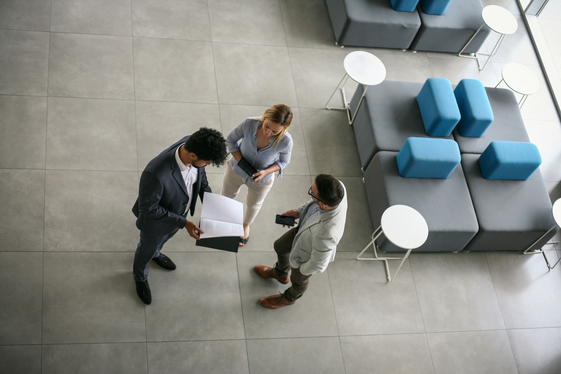 Three people in business attire standing on a tiled floor, reviewing documents near modern seating.