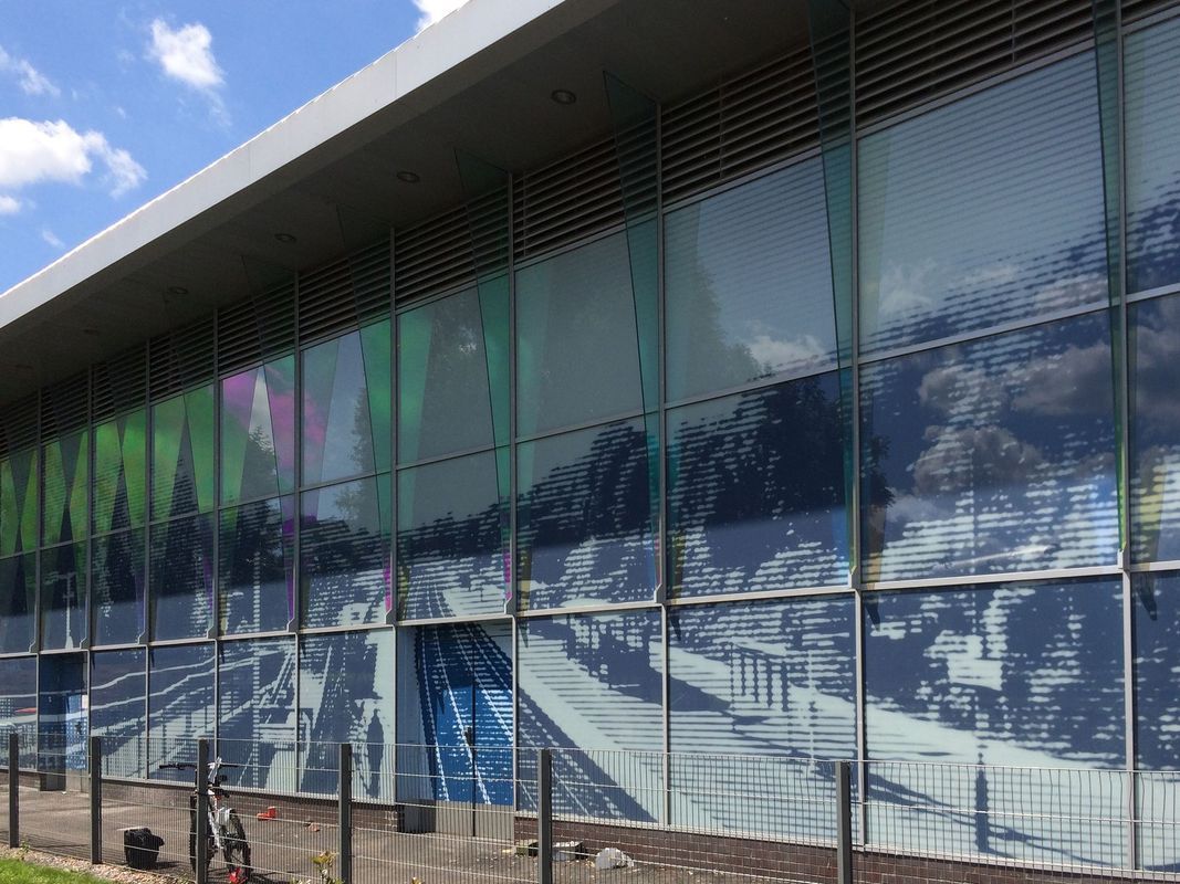 Large glass facade reflecting the sky and surroundings, featuring visible structural beams and an adjacent walkway.