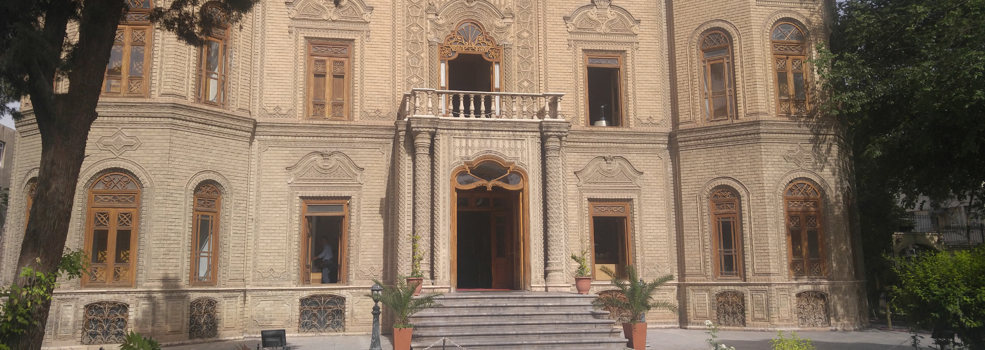 Ornate, beige building with multiple windows and a central entrance with stairs. Green trees frame the sides.