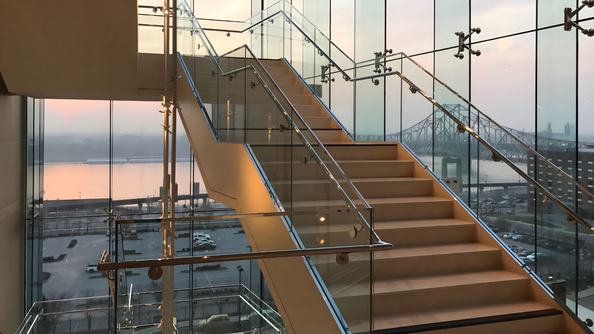 Staircase with glass railing and exterior view of water and city at sunset.