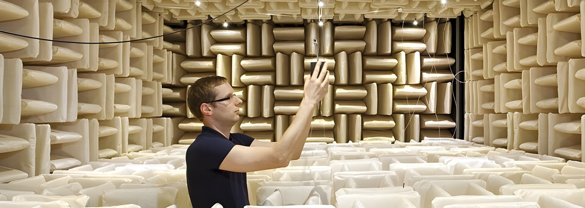 Man in anechoic chamber, holding a device. Beige sound-absorbing panels cover walls and ceiling.