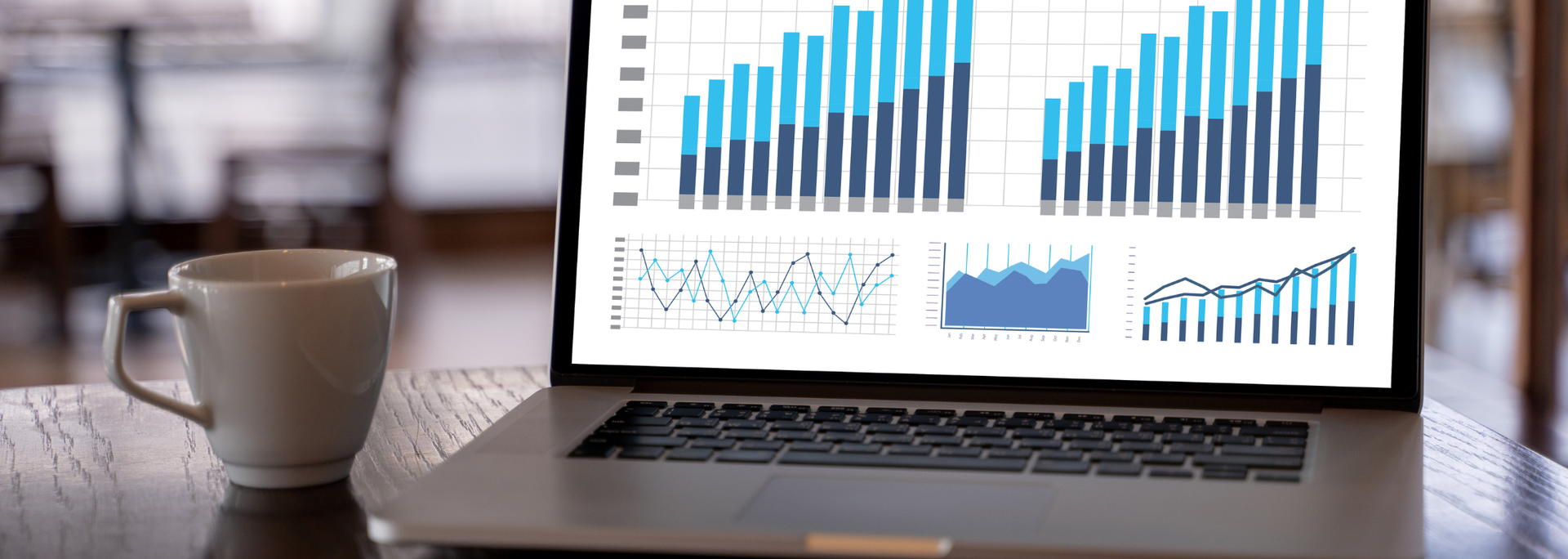 Laptop displaying financial charts, with a coffee cup on a wooden table. Blurred background of a cafe.