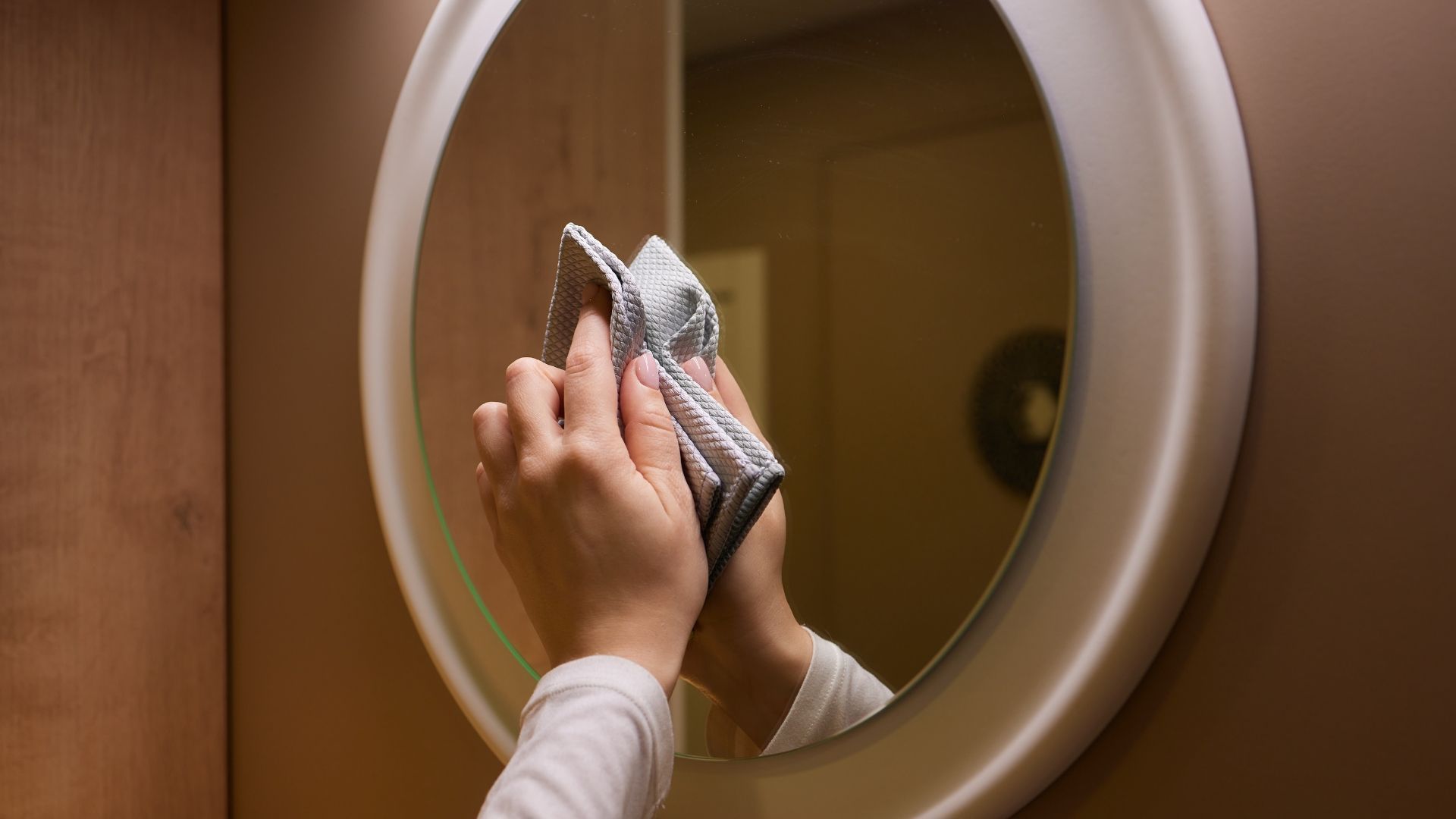 Hands wiping a round mirror with a gray cloth in a brown-walled room.