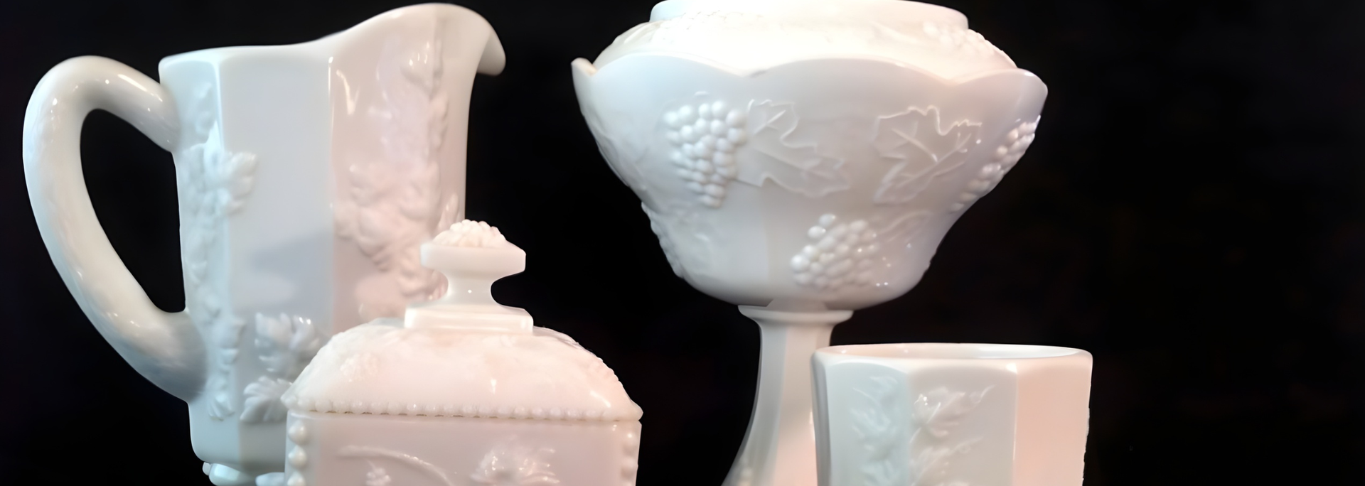 White glass pitcher, covered dish, pedestal bowl, and a small square container. Black background.
