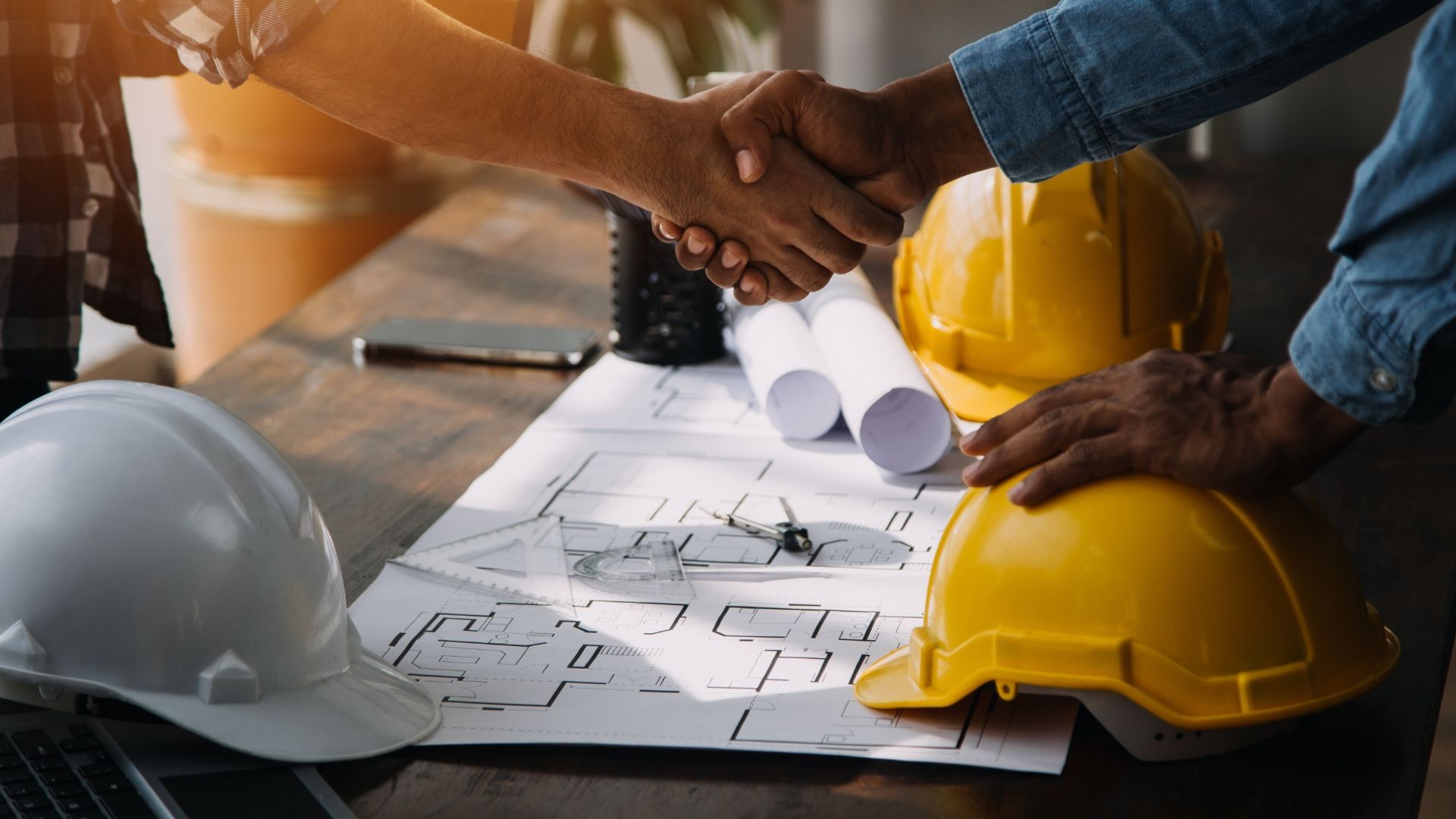 Two people shaking hands over blueprints and hard hats on a desk.