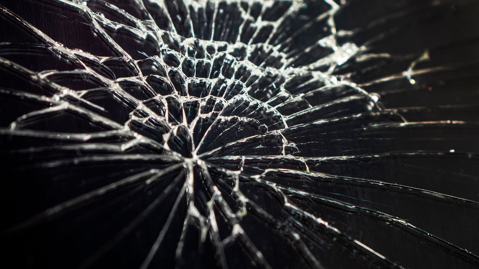 Close-up of a shattered glass screen with a web-like pattern of cracks radiating outward on a dark surface.