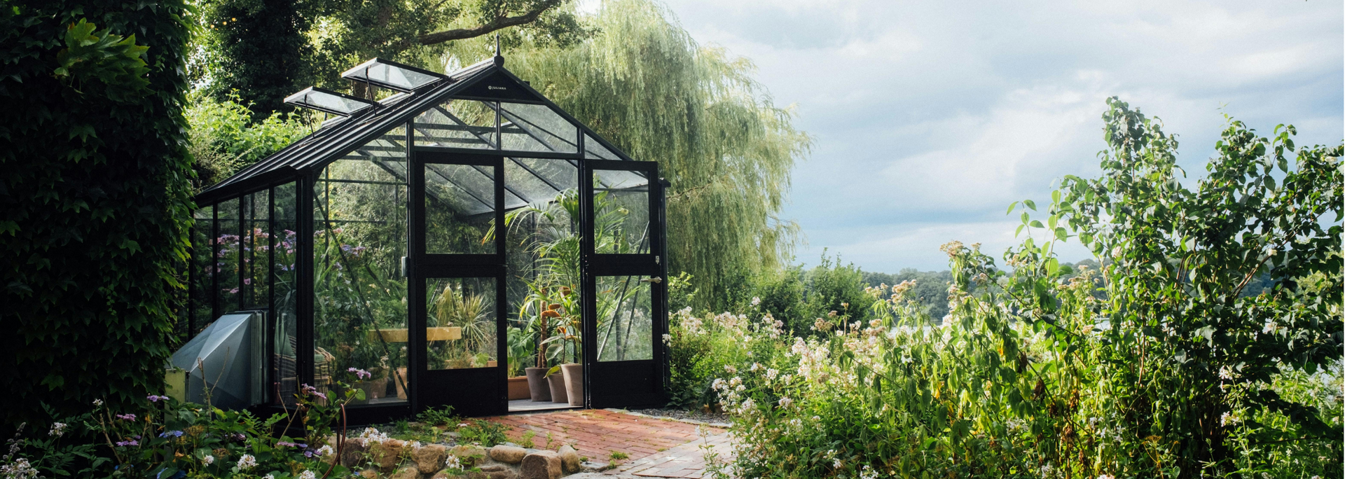 Greenhouse with open doors surrounded by lush greenery under a cloudy sky.