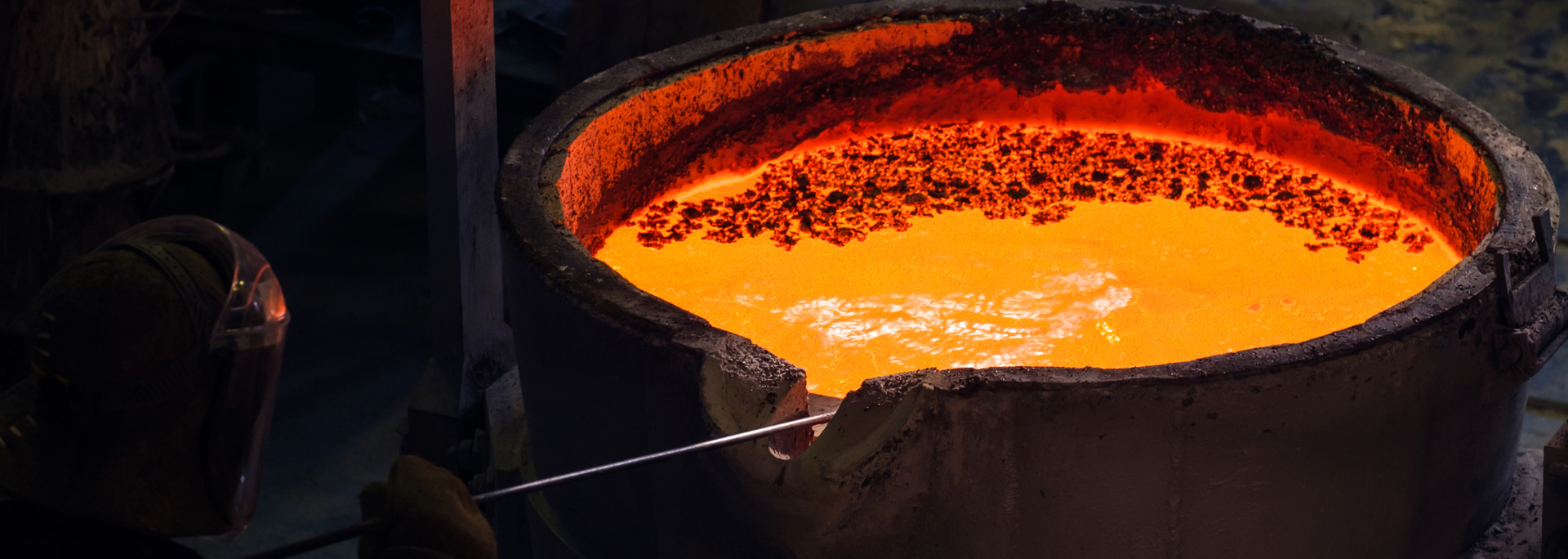 Molten metal in a crucible glows orange. A person with a protective helmet reaches in with a long metal rod.