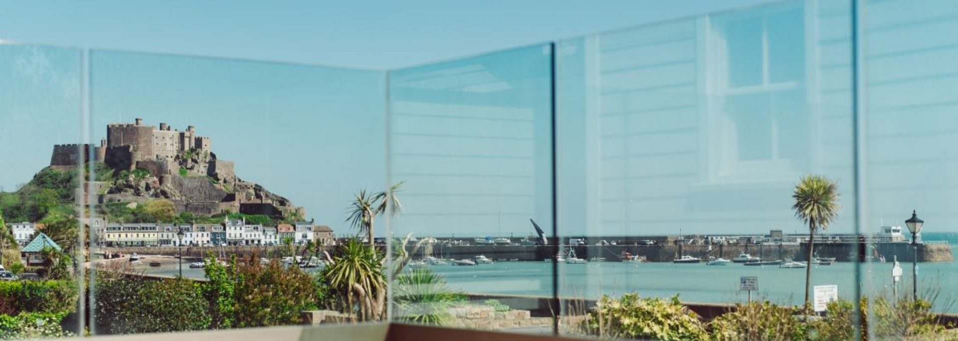 View of a castle on a hill overlooking the sea and harbor, framed by glass panels.