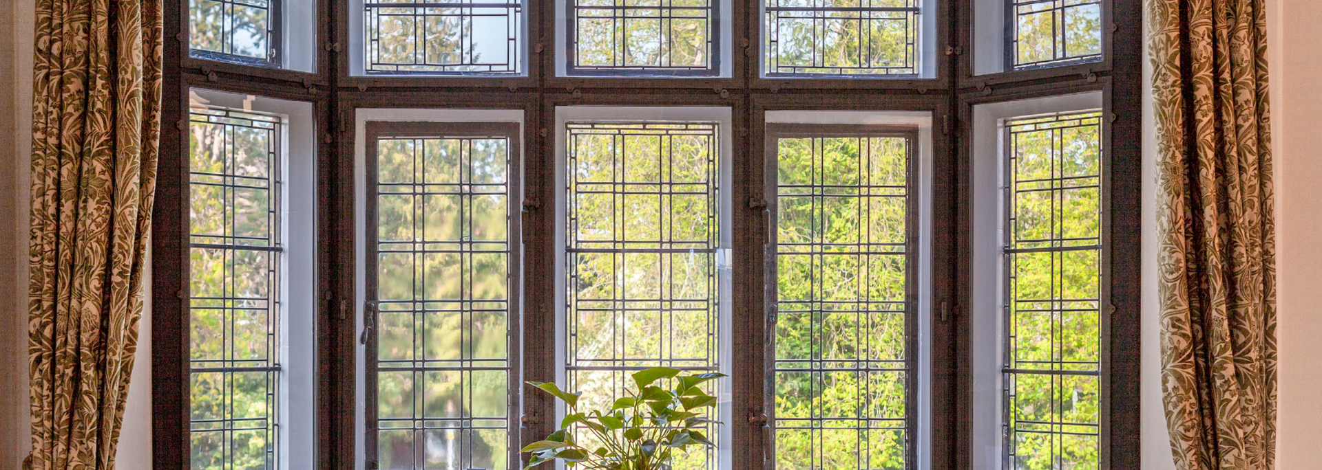 Large bay window with multiple panes, brown frames, and patterned curtains. Bright green trees visible through the glass.