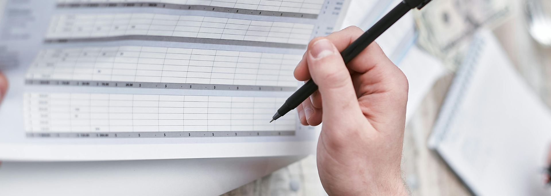 Hand holding a pen, writing on a document filled with rows and columns, possibly financial paperwork.