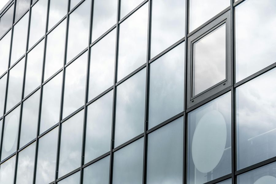 Glass building exterior with single open window, reflecting a cloudy sky.