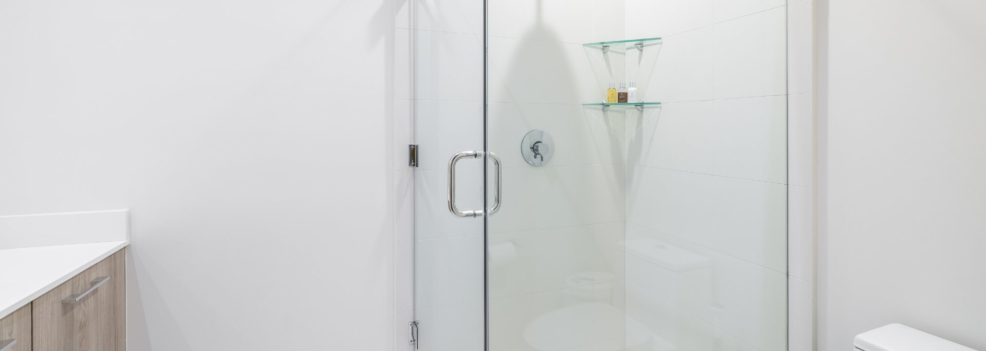 A glass shower with chrome handles and a shelf with bottles, set against a white wall and a light-colored vanity.