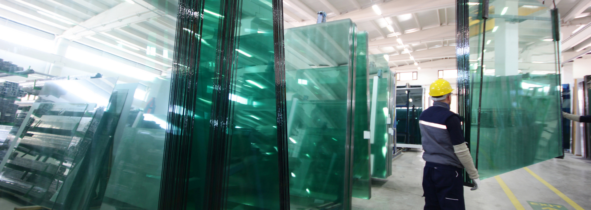 Worker in a factory wearing a yellow hard hat handles large sheets of glass. Warehouse setting.